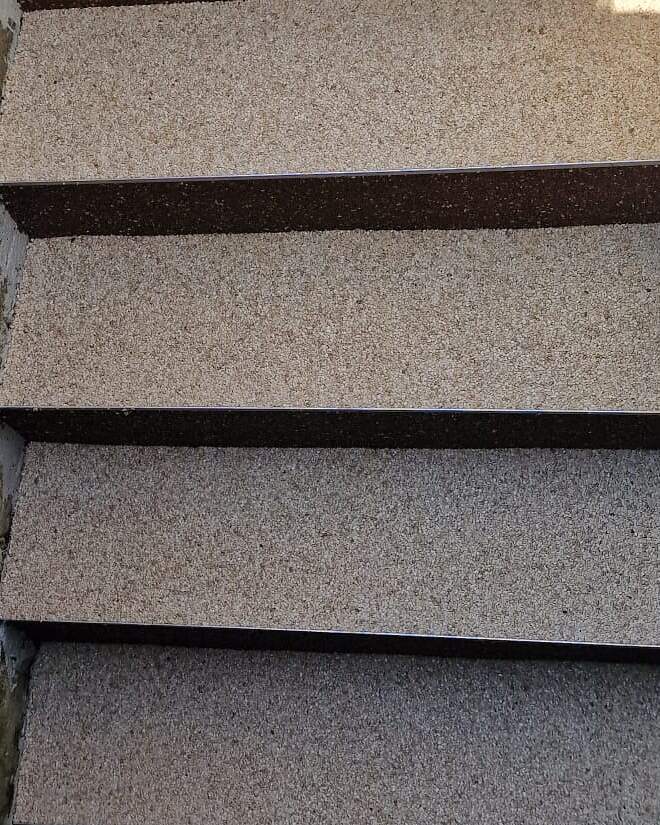 Detailaufnahme einer mit Steinteppich beschichteten Außentreppe: helle Trittstufen mit dunkelbraun abgesetzten Setzstufen und Aluminium-Kantenprofil.
